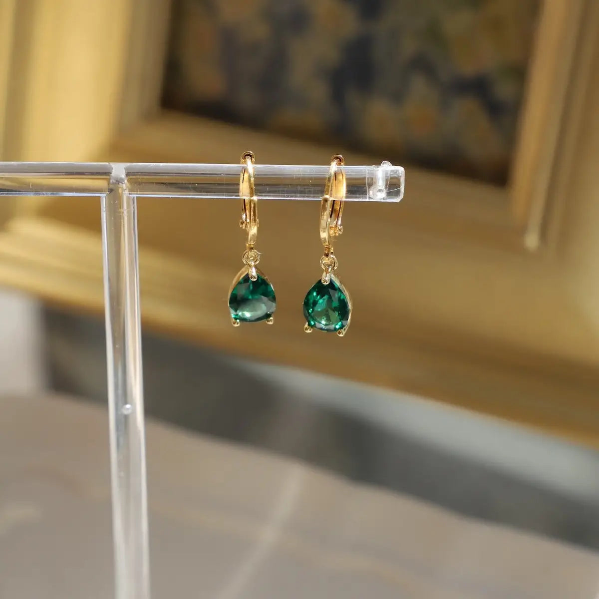 Gold earrings with green gemstones on a clear stand against a blurred background