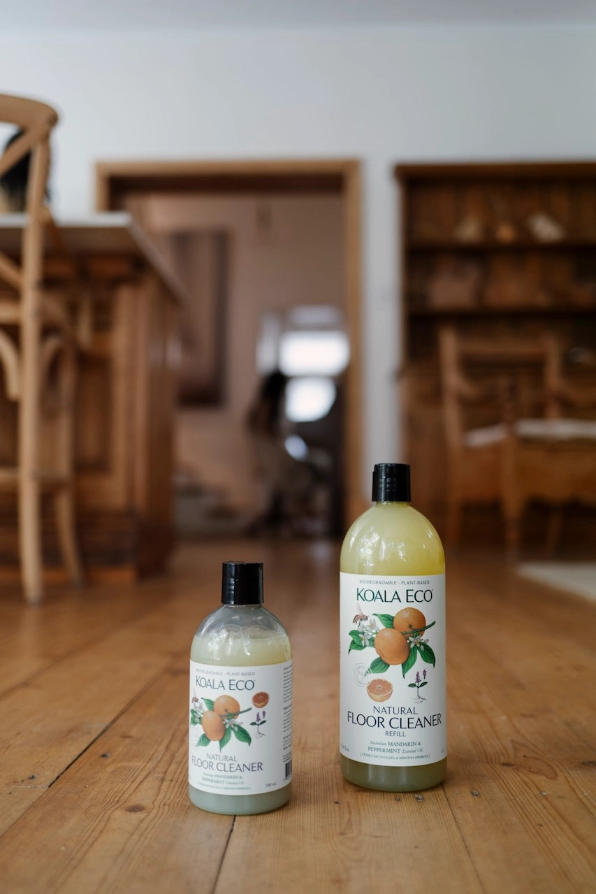 Two bottles of Noala Eco cleaning products on a wooden floor with a blurred room background.