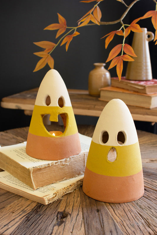 Two ceramic lanterns shaped like candy corn with smiling faces, placed on a wooden surface with a branch in the background and books in the foreground.