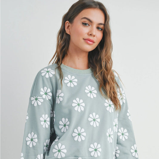 A woman wearing a green round neck sweatshirt with daisy embroidery, paired with white denim shorts.