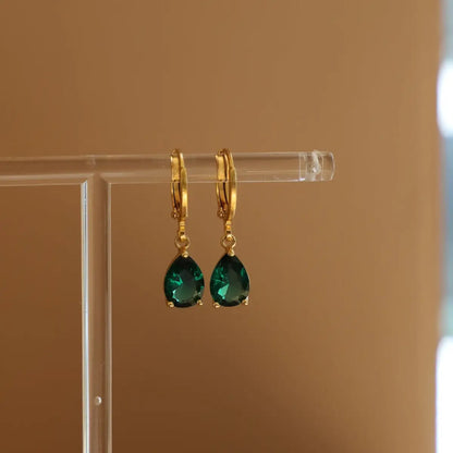 Gold hoop earrings with green teardrop gemstones on a clear stand against a beige background