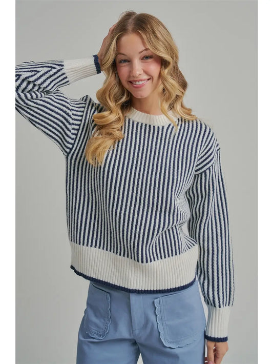 A woman wearing a navy and cream vertically striped knit sweater with ribbed cuffs and hem, paired with light blue jeans, against a solid gray studio background.