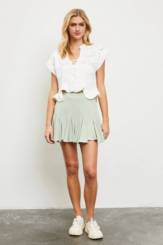 A woman standing in a studio setting wearing a light green flared mini skort with a white, short-sleeved, eyelet-top. She has paired the skort with white sneakers and is posing with a neutral expression.