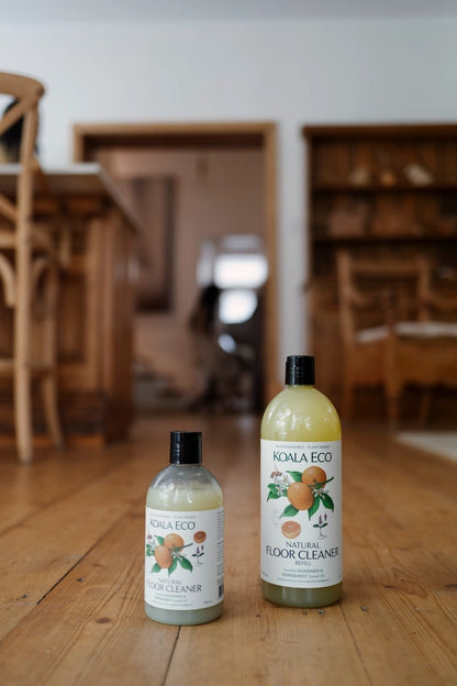 Two bottles of Noala Eco cleaning products on a wooden floor with a blurred room background.