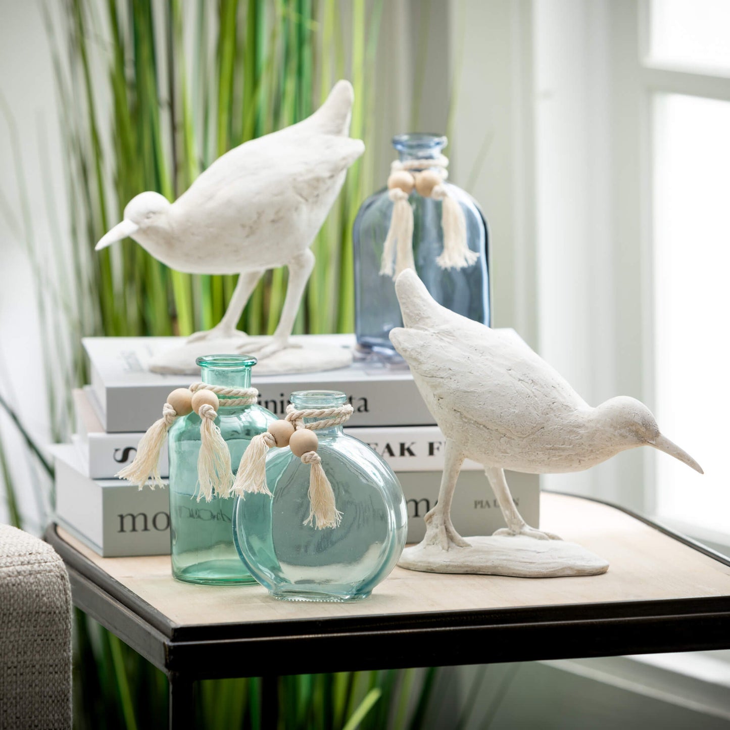 A pair of white resin sea bird figurines with a weathered finish, displayed on a table alongside glass bottles and decorative tassels.
