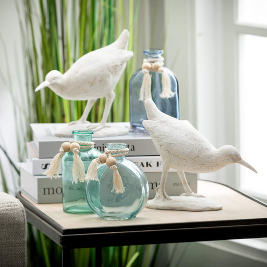 A pair of white resin sea bird figurines with a weathered finish, displayed on a table alongside glass bottles and decorative tassels.