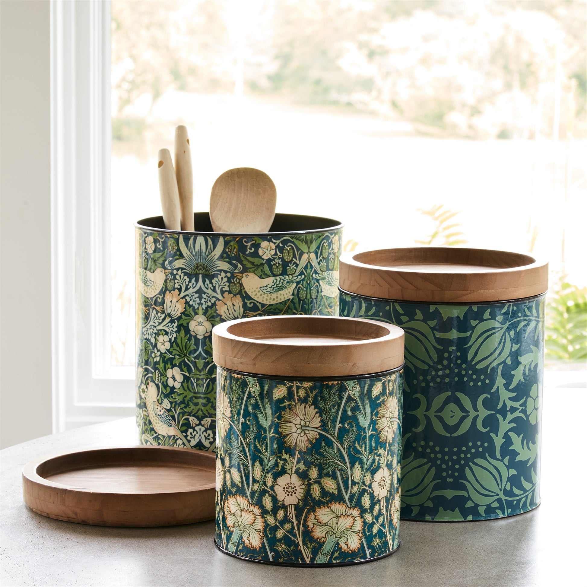 A set of three storage canisters in varying sizes with wood lids, adorned with a green floral pattern, displayed on a kitchen counter.